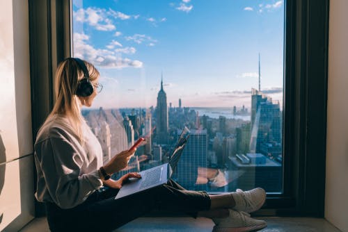Blonde woman looking out a window on the skyline listening to podcasts.